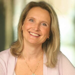 Foto zeigt blonde, lachende Frau an einem Tisch lehnend.